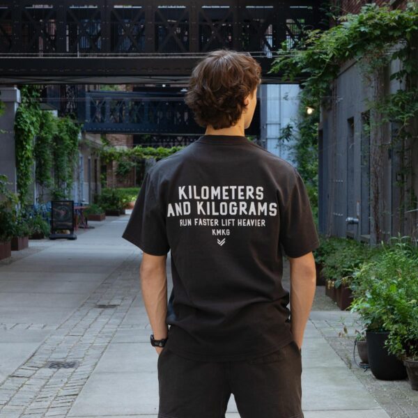 Model draagt de zwarte KMKG Tee met opdruk 'kilometers and kilograms', casual look in een stedelijke setting