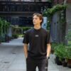 Model draagt de zwarte KMKG Tee met KMKG logo, casual look in een stedelijke setting, voorkant tank