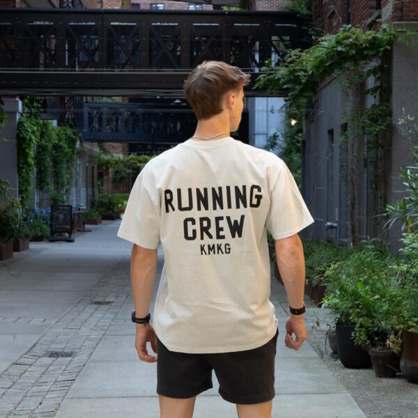 Model draagt de witte KMKG Tee met opdruk 'running crew', casual look in een stedelijke setting