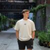 Model draagt de witte KMKG Tee met het KMKG logo als opdruk, voorkant tee, casual look in een stedelijke setting