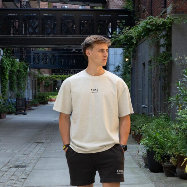 Model draagt de witte KMKG Tee met het KMKG logo als opdruk, voorkant tee, casual look in een stedelijke setting